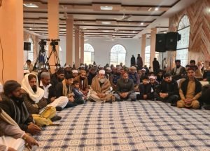 Worshippers seated in rows on the carpet of a mosque hall before congregational prayer