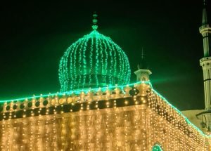 A green-domed shrine illuminated at night during an Islamic commemoration